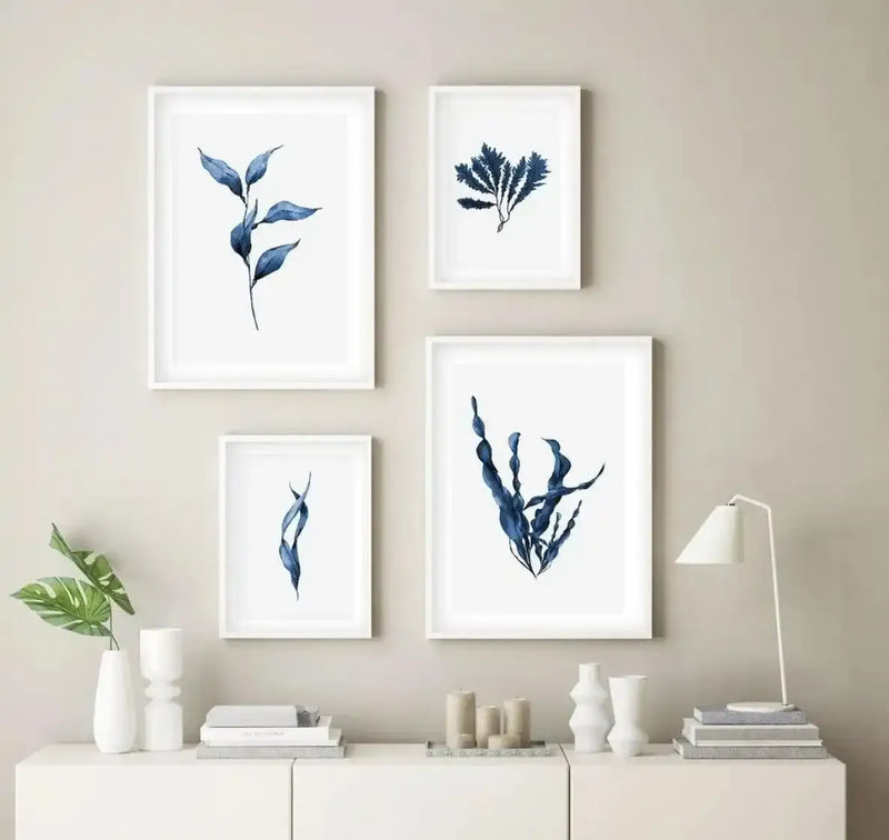 Lifestyle shot of four framed art prints displayed above a white console table in a minimalist living room. The prints feature various blue sea kelp and botanical elements on a white background, with white frames. The console table is adorned with white vases, books, candles, and a white table lamp.