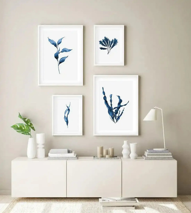 Lifestyle shot of a minimalist living room featuring a white console table with five framed art prints displayed above it on a light beige wall. The prints feature various blue kelp and seaweed illustrations in different shades of deep blue and indigo, each with a white border and white frame. The console table is adorned with white vases, candles, and books, and a white table lamp sits on the right side.