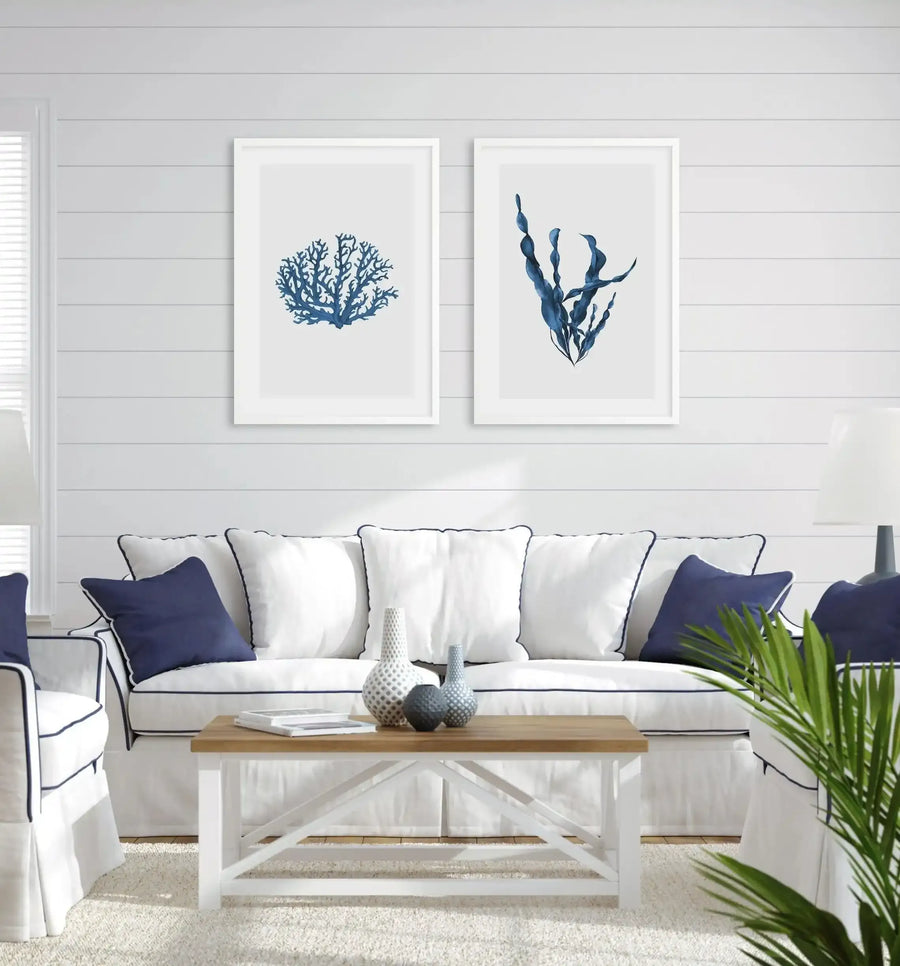 Lifestyle shot of a living room with two framed art prints displayed above a white sofa. The left print features a blue coral illustration, and the right print features a blue sea kelp illustration. Both prints have a white border and a white frame. The room has white shiplap walls, a white sofa with navy blue piping and cushions, a light wood coffee table with white legs, and a cream rug.
