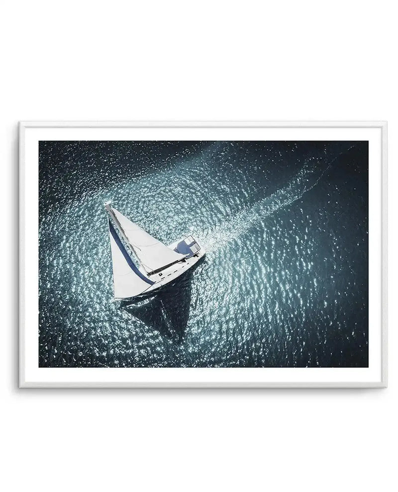 Framed aerial photograph of a sailboat on a shimmering dark blue ocean, with a white frame and white border. The sailboat, with white sails and blue accents, casts a dark shadow on the water and leaves a trail of white foam behind it. The water reflects sunlight, creating bright, sparkling patches.