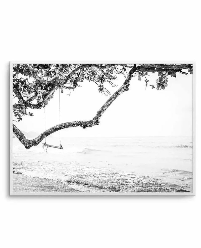 Black and white photograph framed in light gray showing a gnarled tree branch extending horizontally over a beach. The branch has detailed texture and scattered foliage at its top and ends. A rope swing hangs from the branch with simple knots. Below, a sandy beach meets calm ocean waters with gentle waves. The composition captures a serene, minimalist coastal scene with high contrast between the dark tree and light sky. The print includes a white border within the gray frame.