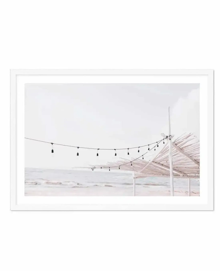 Framed photograph of a serene beach scene with a string of lights and a white thatched structure, displayed in a white frame with a white border. The composition features gentle waves rolling onto a sandy shore under a soft, overcast sky, creating a light and airy mood.