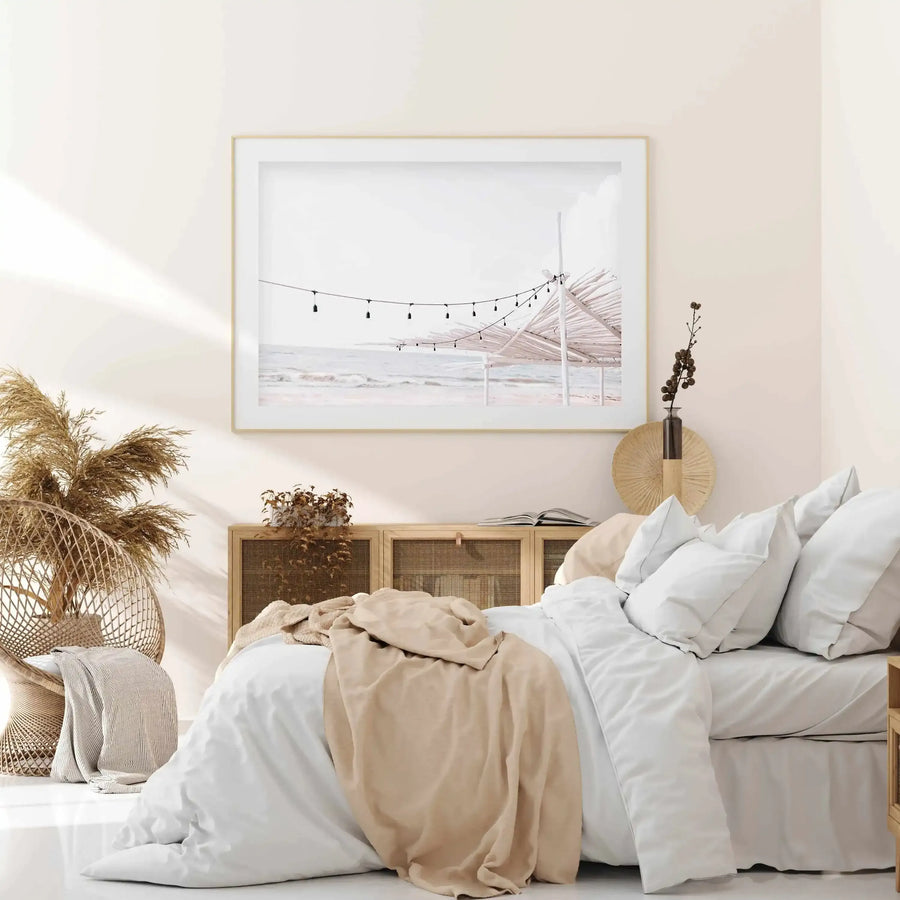 Lifestyle shot of a bedroom featuring a large framed art print above a bed. The print depicts a serene beach scene with a string of lights hanging above a thatched umbrella, with gentle waves and a soft, overcast sky. The art print is displayed in a natural wood frame with a white border. The bedroom features a bed with white bedding and a beige throw, a wicker chair with dried pampas grass, and a wooden cabinet with cane detailing.