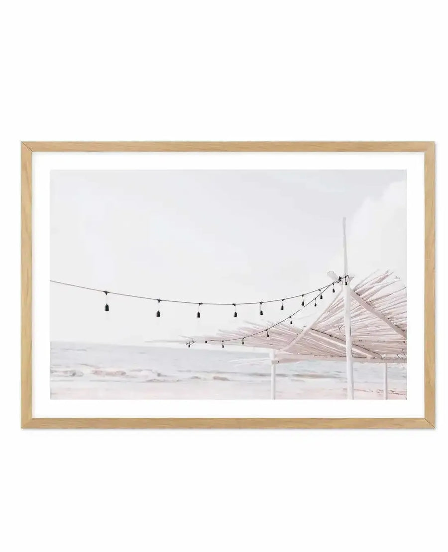 Framed photography art print of a serene beach scene with a natural wood frame and white border. The composition features a string of black Edison-style lights strung across the frame, leading to a light-colored, thatched-roof structure on the right, with the gentle, pale blue ocean and a soft white sky in the background.