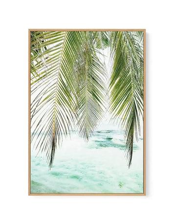 Framed canvas art print depicting a tropical beach scene viewed through hanging palm fronds. Green and sage-toned palm leaves with fine linear details frame the composition on both sides, creating a natural window. Beyond the fronds, turquoise shallow water and pale sand stretch toward the horizon beneath a soft, overcast sky. The artwork has a serene, minimalist quality with a soft, watercolor-like finish. The canvas is mounted in a natural wood frame with a light tan finish.