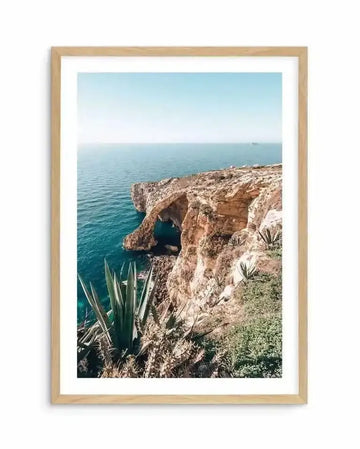 Framed photograph of a coastal scene in Malta with a natural wood frame and white border. The image captures a rugged cliff face with natural arch formations, meeting the turquoise-blue Mediterranean Sea under a clear, bright sky. Lush green agave plants and other coastal vegetation grow on the sun-drenched cliffs, with a distant ship visible on the horizon.