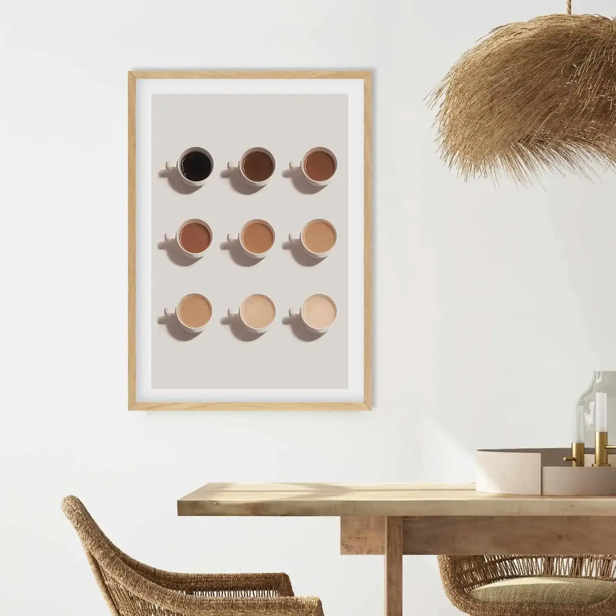 Framed art print featuring nine cups of coffee, arranged in three rows of three, displayed on a light grey background, with a natural wood frame and white border. The coffee shades range from dark black in the top left to a very pale cream in the bottom right, creating a gradient effect. The print is hung on a white wall above a light wood dining table with a woven chair in the foreground and a straw-like pendant light fixture hanging above.
