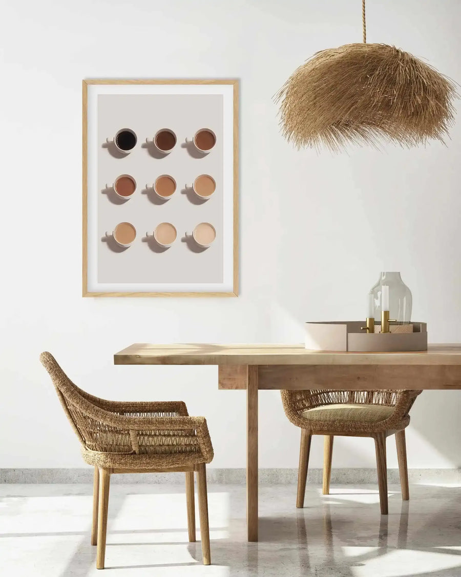 Lifestyle shot of a dining room with a framed art print displayed on a white wall. The print features nine cups of coffee in varying shades from dark black to pale cream, arranged in a 3x3 grid on a light grey background, with a natural wood frame and white border. The room includes a light wood dining table with two woven wicker chairs, a large straw pendant light hanging from the ceiling, and a decorative tray with a glass vase and two candles on the table.