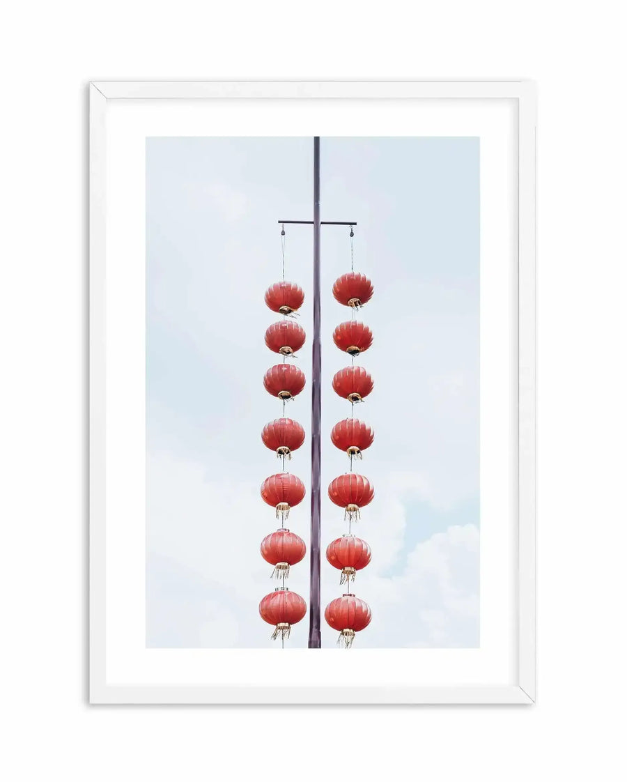 Shanghai Days Art Print displayed in a white frame with white border. The artwork is a vertical color photograph featuring two parallel strands of red paper lanterns hanging from a horizontal dark pole against a light blue cloudy sky. Each strand contains eight round lanterns with ribbed texture and small tassel details at the base. The lanterns are symmetrically arranged, creating a balanced, minimalist composition with clean lines and soft, muted tones.
