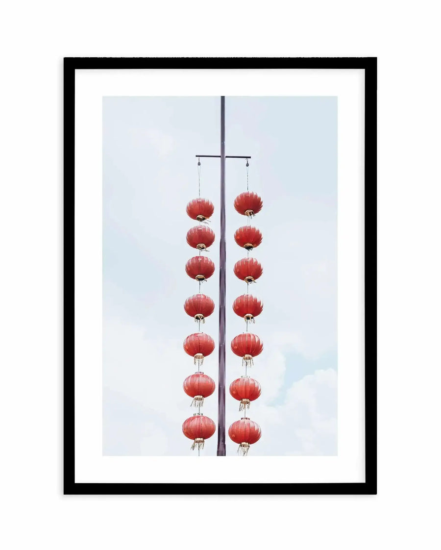 Framed art print featuring a photograph of two vertical strings of red paper lanterns hanging from a horizontal black pole against a light blue cloudy sky. Each string contains eight rounded, ribbed lanterns with small tassels at the bottom, arranged symmetrically on either side of the central pole. The photograph has a minimalist, balanced composition. The print is mounted in a black frame with a white border.