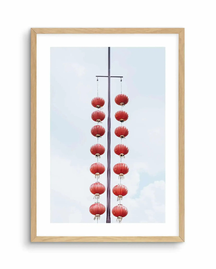 Shanghai Days Art Print framed in light natural wood. The artwork features a photograph of two vertical strands of red fabric lanterns hanging from a dark metal pole against a pale blue sky with white clouds. Each strand contains eight round lanterns with ribbed texture and gold-toned metal fittings at the base. The composition is symmetrical and minimalist, with the lanterns centered against the soft overcast sky. The print is mounted with a white border.