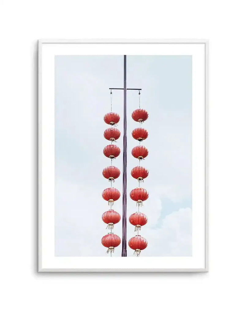 Framed art print featuring a vertical photograph of red paper lanterns suspended from a dark pole against a pale blue sky. Fourteen round lanterns with ribbed texture and gold-toned metal fixtures hang in two symmetrical columns of seven on either side of the central pole. The lanterns appear evenly spaced, creating a balanced, minimalist composition. The artwork is presented in a silver-toned frame with a white border.