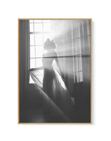 Black and white framed canvas art print featuring a silhouetted figure of a woman in flowing fabric standing by a large gridded window with sheer curtains. Dramatic backlighting creates luminous rays streaming through the window, casting the figure in shadow. The composition emphasizes contrast between bright light and dark silhouette. Natural wood frame with vertical portrait orientation. Soft, ethereal mood with high-contrast monochromatic photography style.