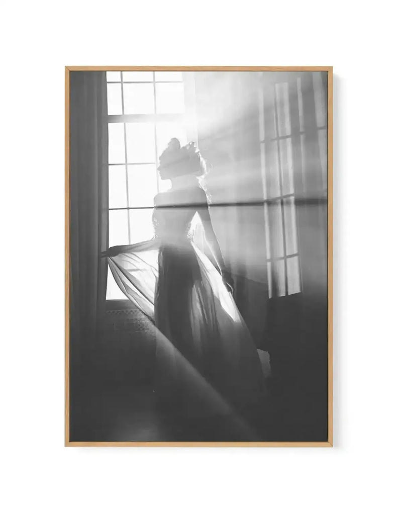Black and white framed canvas art print featuring a silhouetted figure of a woman in flowing fabric standing by a large gridded window with sheer curtains. Dramatic backlighting creates luminous rays streaming through the window, casting the figure in shadow. The composition emphasizes contrast between bright light and dark silhouette. Natural wood frame with vertical portrait orientation. Soft, ethereal mood with high-contrast monochromatic photography style.