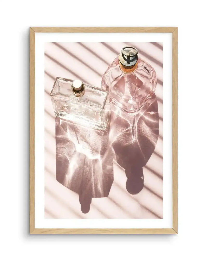 Framed art print featuring a top-down photograph of two empty glass perfume bottles on a light pink surface, with strong shadows and light reflections creating a striped pattern. The rectangular bottle is on the left, and the rounded square bottle is on the right, both casting long, dark shadows. The light creates shimmering patterns on the surface around the bottles, and the overall mood is bright and airy. The print is displayed in a natural oak frame with a white border.