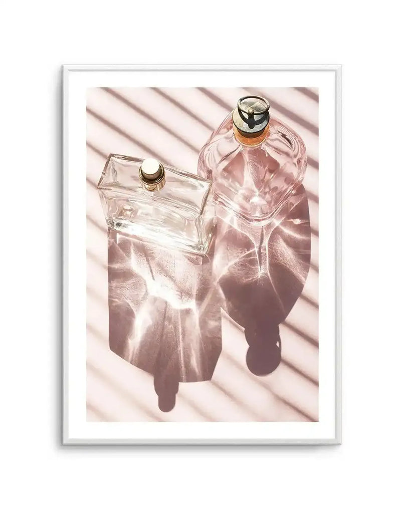 Framed photograph of two perfume bottles on a pale pink surface with horizontal shadows and light refractions. The left bottle is clear glass and rectangular with a gold and white cap. The right bottle is blush pink glass and heart-shaped with a gold and black cap. Both bottles cast long, dark shadows and create shimmering light patterns on the surface, with a white frame and border.