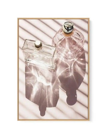 Framed canvas art print featuring two glass perfume bottles photographed from above against a white background with diagonal shadow patterns. The left bottle is a clear rectangular flacon with a gold-trimmed stopper, while the right bottle is a rounded shape filled with pale rose-tinted liquid, also topped with a gold and black cap. Soft shadows cast across the composition create depth and dimension. The artwork is mounted in a natural wood frame with a white border.