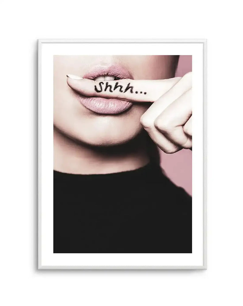 "Framed photographic art print of a woman's lower face with a finger pressed to her lips, displayed in a white frame with a white border. The woman has light pink lipstick and a black top, with the word 'Shhh...' written in black on her finger. The background is a soft pink."