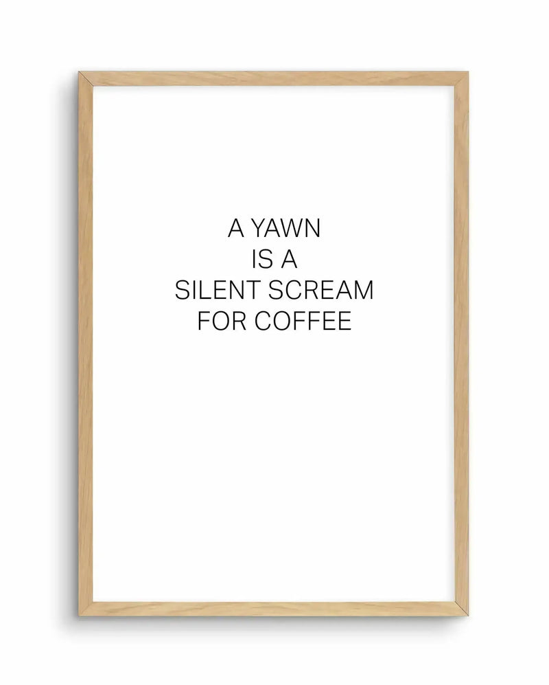 Framed art print with natural wood frame displaying black text on white background. Text reads "A YAWN IS A SILENT SCREAM FOR COFFEE" centered on the page in a clean, sans-serif typeface. The minimalist design features simple black lettering with generous white space, creating a humorous statement about caffeine dependency. The light wood frame has a subtle grain texture and sits flush against the white border of the print.