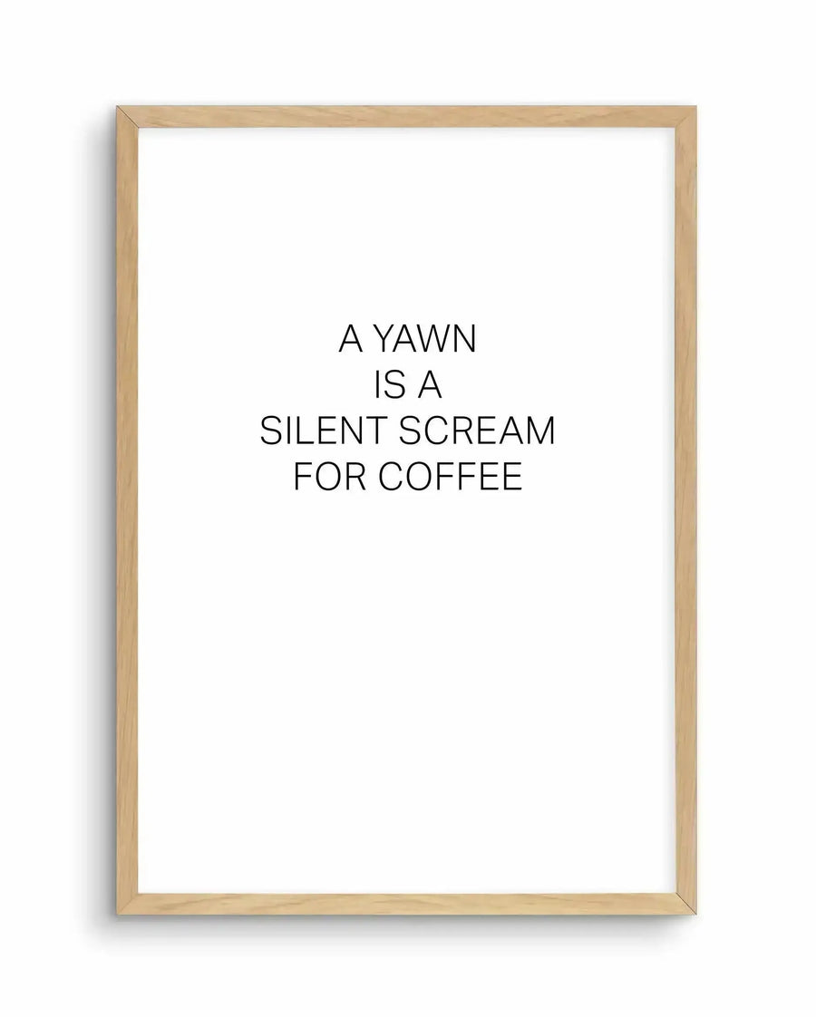 Framed art print with natural wood frame displaying black text on white background. Text reads "A YAWN IS A SILENT SCREAM FOR COFFEE" centered on the page in a clean, sans-serif typeface. The minimalist design features simple black lettering with generous white space, creating a humorous statement about caffeine dependency. The light wood frame has a subtle grain texture and sits flush against the white border of the print.