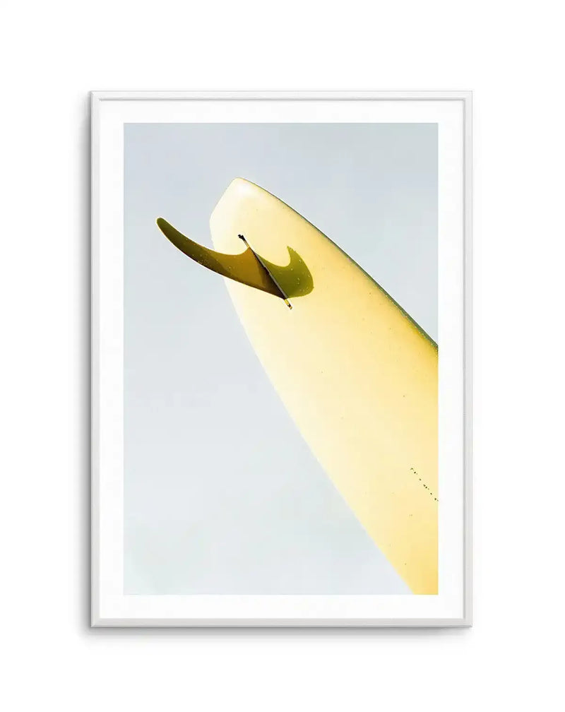 Art print titled "Single Fin Burnt Lemon" by Mario Stefanelli displayed in a silver frame with white border. The artwork features a minimalist composition of a single surfboard fin against a pale blue-grey background. The fin is rendered in deep olive and mustard yellow tones, showing dimensional shading and a glossy finish. The surfboard appears in pale yellow with subtle gradation, angled diagonally across the frame. The illustration style is clean and contemporary with a calm, understated aesthetic.