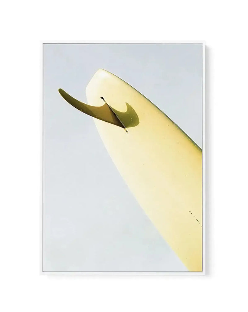 Framed canvas print featuring a close-up, low-angle photograph of a surfboard fin and tail against a bright, hazy sky. The surfboard is a pale yellow, almost cream color, with a subtle sheen, and the fin is a darker, olive-green hue, casting a soft shadow on the board. The composition is diagonal, with the surfboard extending from the lower left to the upper right, and the fin is prominently featured in the upper left quadrant. The image is framed in a simple white frame.