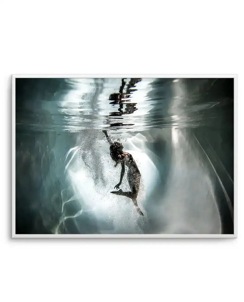 Framed photography art print of a woman underwater, with a white frame and white border. The woman is suspended in the water, with her head tilted back and arms reaching upwards, surrounded by bubbles and illuminated by a bright light source from below. The water is a cool blue-grey, with reflections visible on the surface.