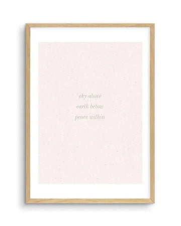 Art print featuring the text "sky above earth below peace within" in a delicate, light green serif font, centered on a soft pink background with a subtle speckled texture, displayed in a natural oak frame with white border.