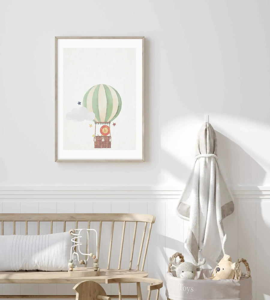 Lifestyle shot of a child's room featuring a framed art print above a wooden bench. The print depicts a hot air balloon with green and cream stripes, carrying a lion in a basket, surrounded by clouds and stars. The room has white paneled walls, a white cushion on the bench, a wooden toy, a grey hooded towel on a hook, and a "Toys" basket with stuffed animals.