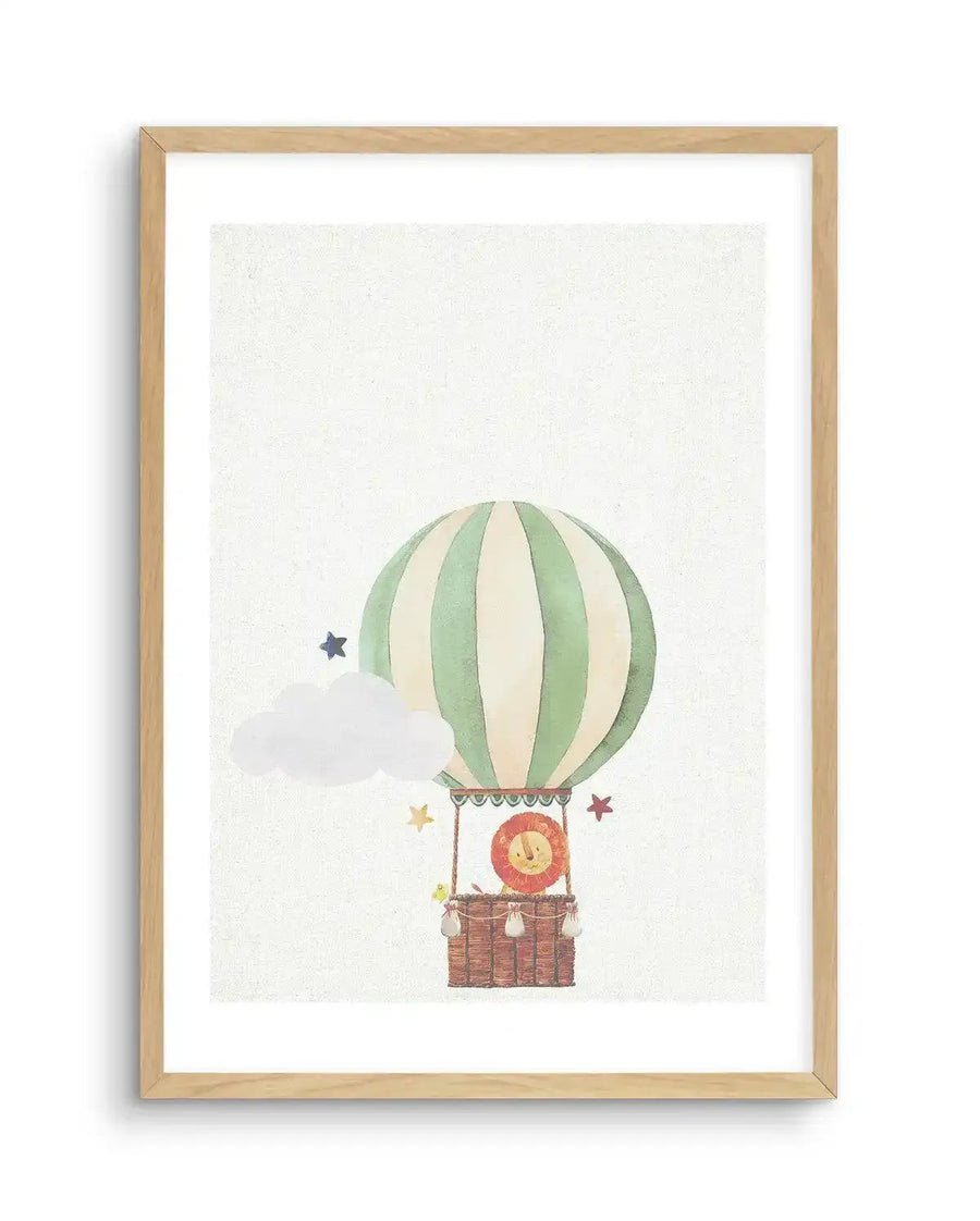 Watercolour art print featuring a hot air balloon with a lion in its basket, printed on fine art paper and displayed in a natural oak frame with white border. The balloon is striped in pale green and cream, with a brown woven basket below. A smiling lion with a red mane peeks out from the basket, accompanied by a small yellow bird. A white cloud and several stars in blue and yellow float around the balloon.