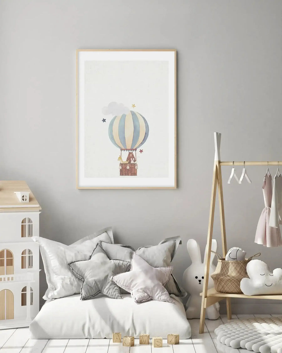 Lifestyle shot of a child's bedroom featuring a framed art print above a low bed with white bedding and star-shaped pillows. The art print depicts a hot air balloon with pale blue and cream stripes, carrying a bear, a deer, and a rabbit in its wicker basket, surrounded by soft grey clouds and gold and red stars on a white background. The room includes a white dollhouse, a wooden clothes rack with children's clothes, a large white bunny toy, and wooden alphabet blocks on the white floorboards.