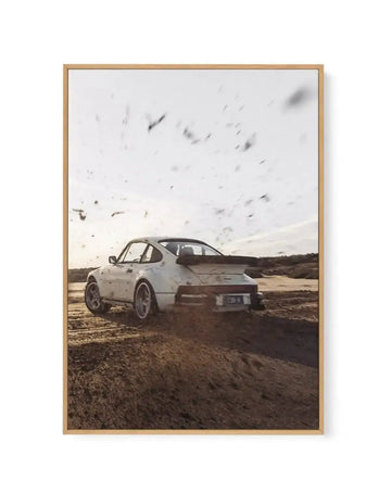 Framed canvas print of a white Porsche 911 Slantnose driving through a sandy, desert-like landscape, kicking up dirt and dust into the bright, hazy sky. The car is seen from the rear, slightly angled, with a large spoiler and a license plate that reads "139 H". The print is displayed in a natural oak frame.