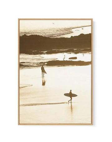 Framed sepia-toned photograph of a coastal beach scene with natural wood frame and white border. The composition features two surfers, silhouetted against the bright sand, walking along wet sand at low tide with gentle waves, scattered rocks, and birds in the distance under a bright sky.