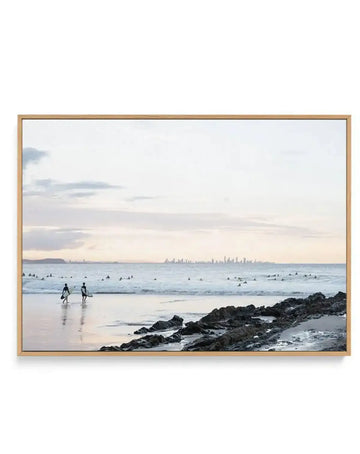 Framed canvas art print featuring a coastal beach scene at dusk with two surfers in wetsuits walking into the water on the left, numerous surfers on boards scattered in the ocean, dark volcanic rocks in the foreground, calm waves, and a distant city skyline visible across the water under a soft grey and white cloudy sky. The photograph has a muted, serene colour palette of pale blues, whites, and greys. The canvas is mounted in a natural wood frame with a white border.