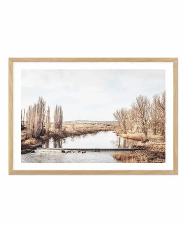 Framed photography art print of a serene river landscape in muted tones, displayed in a natural oak frame with white border. The composition features a calm river with a small weir, flanked by tall, bare trees with light brown foliage on both banks, reflecting in the water. In the background, rolling hills with dry grass stretch under a soft, overcast sky.