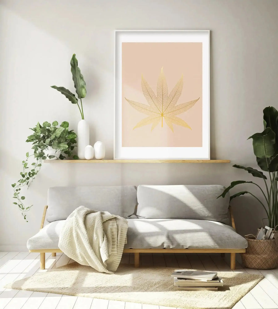 Lifestyle shot of a minimalist living room featuring a large framed art print of a golden cannabis leaf skeleton on a soft peach background, displayed above a light grey sofa. The print has a white frame and white border. The room includes a trailing green plant in a white pot, a tall green plant in a woven basket, and a cream throw blanket draped over the sofa.