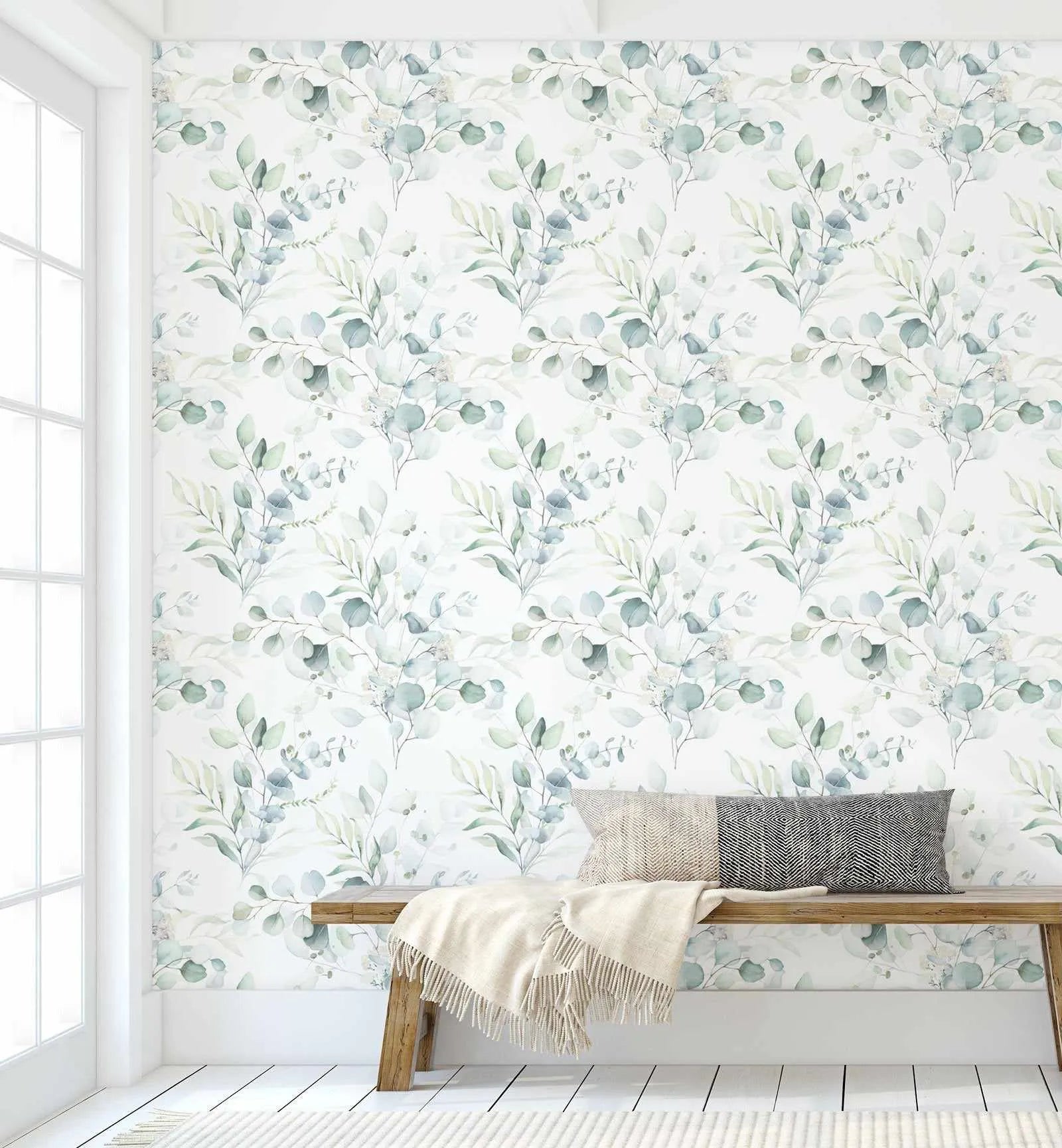 Lifestyle shot featuring Soft Eucalyptus Wallpaper in a bright room with white walls, floor, and trim. The wallpaper features a repeating pattern of hand-painted eucalyptus branches in muted sage green and dusty blue-green on a soft white background. The botanical design has a loose, organic watercolour quality with visible brushstroke texture. A light wood bench with a cream throw blanket and two patterned pillows sits against the wallpapered wall. A large window with white panes is visible on the left.
