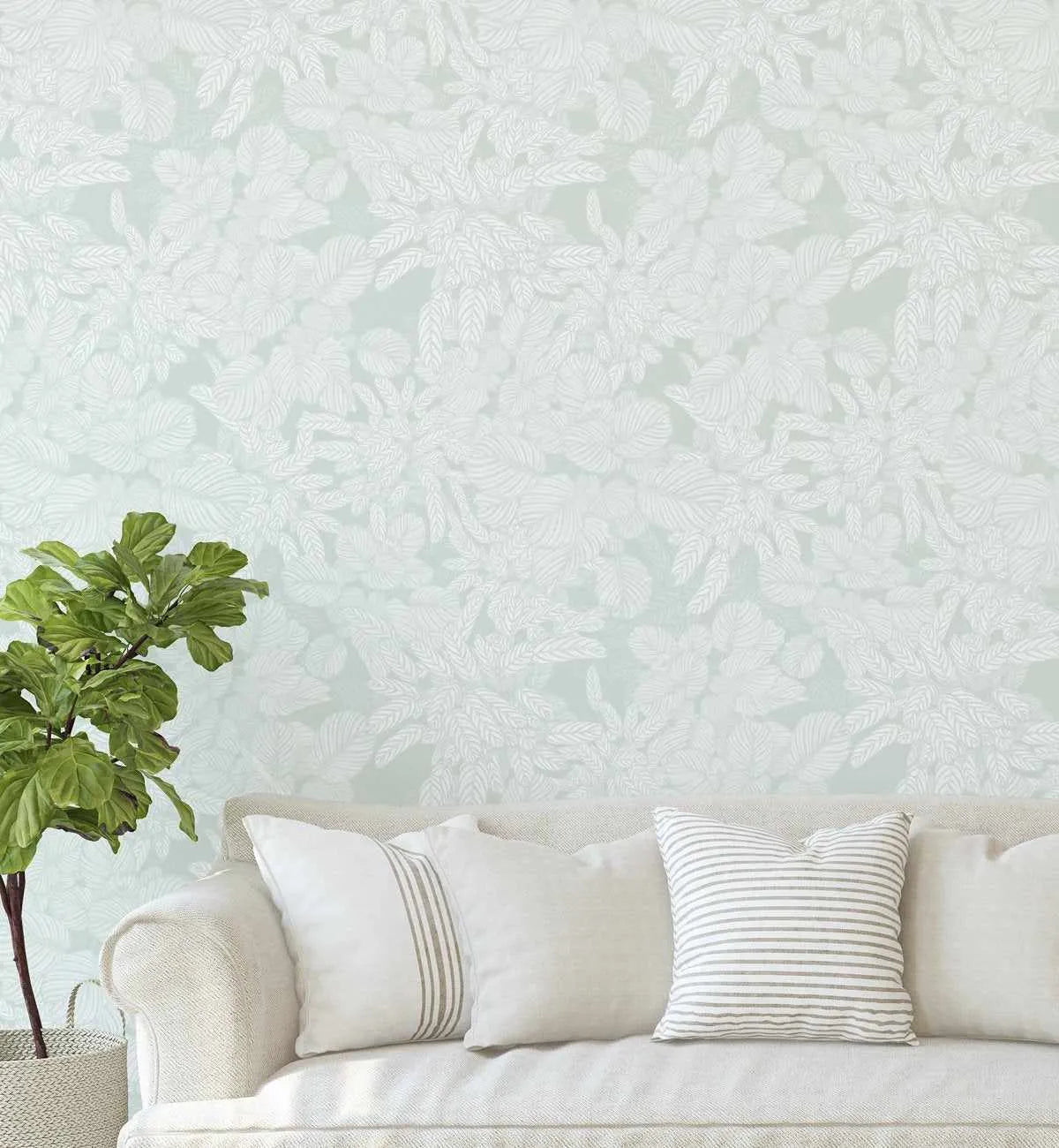Lifestyle shot of a living room featuring Soft Foliage Sage Green Wallpaper. The wallpaper has a repeating pattern of hand-drawn tropical leaves and fronds in an off-white colour against a soft sage green background. A cream-coloured sofa with three white and striped throw pillows is in the foreground, and a potted fiddle-leaf fig plant sits to the left.
