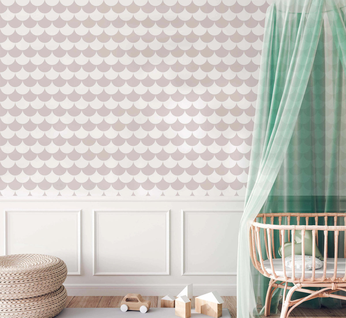Soft Grey Scallops Wallpaper displayed in a nursery room setting. The wallpaper features a repeating pattern of overlapping scallop shapes in soft mauve, dusty rose, and cream tones on a pale background, creating a gentle wave-like texture. The room shows white wainscoting on the lower wall with the patterned wallpaper above, natural wood flooring, a woven round floor cushion, wooden toy blocks, a toy car, and a natural wood crib with sheer sage green canopy draping on the right side.