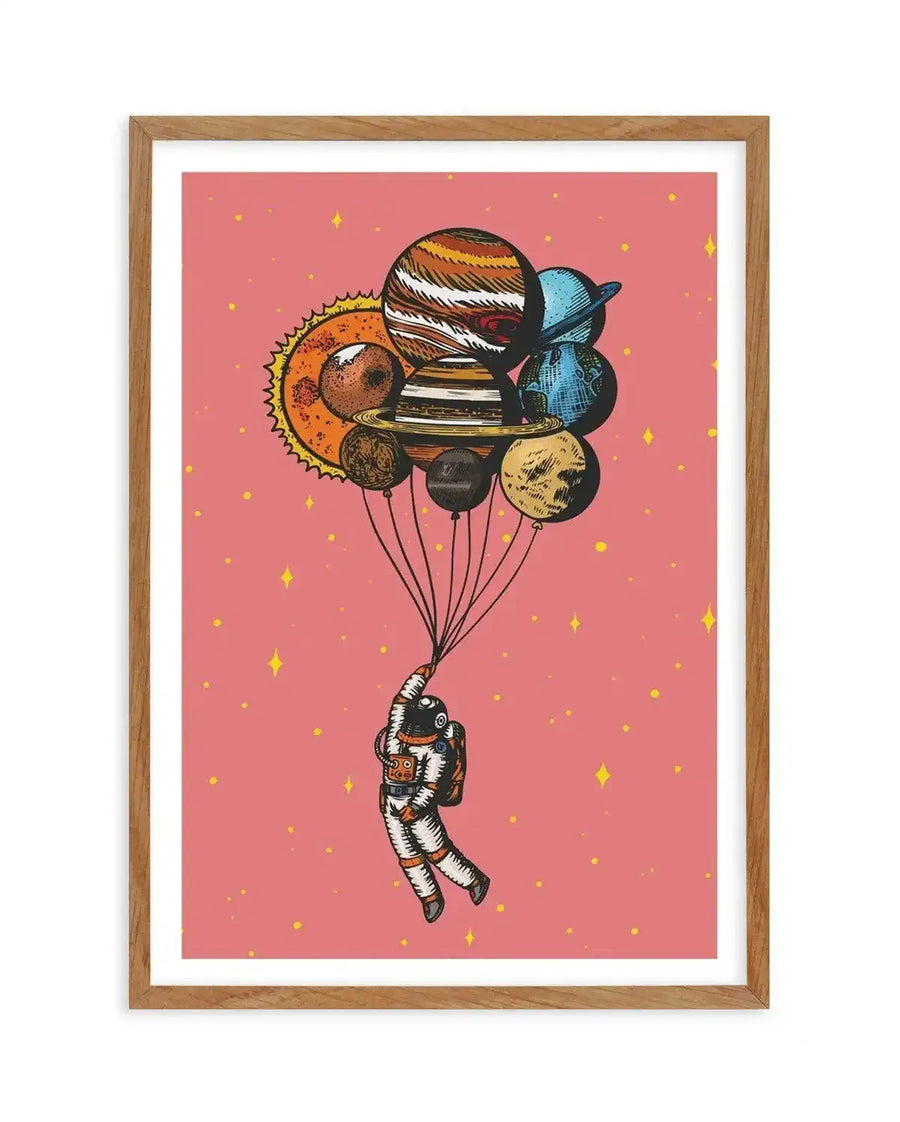 "Whimsical art print featuring an astronaut floating in space, holding onto a cluster of balloons shaped like planets and the sun, against a speckled pink background with yellow stars. The illustration has a hand-drawn, vintage comic book style with visible line work and vibrant colours, displayed in a natural oak frame with white border."