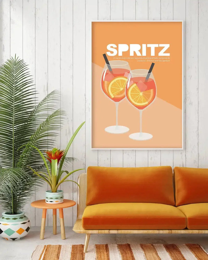 Large framed art print displayed above an orange sofa in a bright living room. The print features two Aperol Spritz cocktails with orange slices and straws on an orange background, with a white frame and white border. The room includes a large green plant in a patterned pot, a smaller plant in a striped pot on a small orange table, and a striped rug.