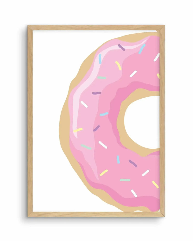 Framed art print featuring an illustration of a pink frosted donut with colorful sprinkles in a natural wood frame. The donut design shows layered pink icing with a golden-tan glazed exterior, a white center hole, and multicolored sprinkles in blue, purple, yellow, white, and green scattered across the frosting. The artwork is rendered in a flat, graphic illustration style with soft shading against a white background and white border within the frame.