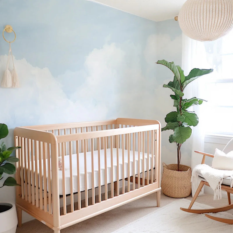 Nursery:
Peel and stick wallpaper featuring a hand-painted cloudy sky mural in soft pastel blue and wispy white, covering the wall behind a light wood crib in a bright nursery. The room includes a large fiddle leaf fig plant in a woven basket, a light wood rocking chair with a cream cushion and throw, and a cream woven pendant light fixture.