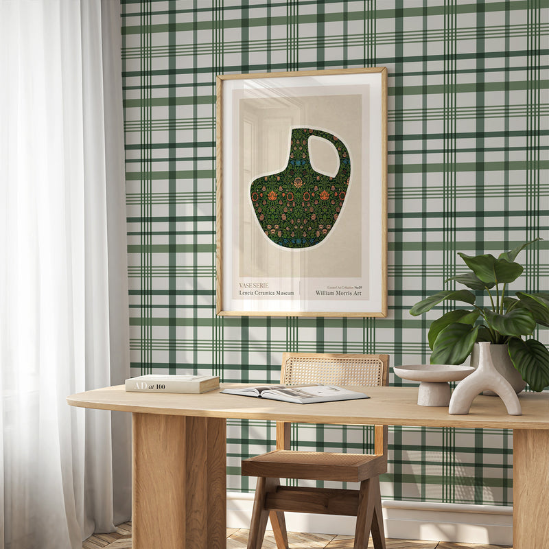 Dark green plaid wallpaper with a woven texture displayed on a bedroom wall. The pattern features a classic tartan design in forest green and cream with intersecting horizontal and vertical stripes creating a check pattern. The wallpaper is shown in a styled interior with a light wood desk, rattan chair, framed botanical vase artwork, potted plant, and white curtains, demonstrating how the wallpaper complements mid-century modern decor.