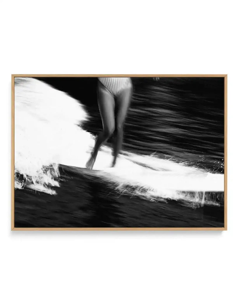 Framed black and white photograph of a surfer riding a wave, printed on canvas and displayed in a natural wood frame. The composition captures the surfer's legs and lower torso in a striped swimsuit, with motion blur on the water creating a dynamic sense of movement.