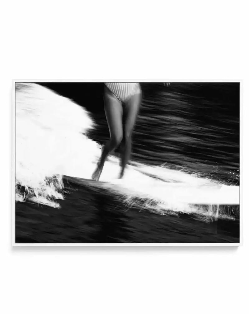 Framed black and white photograph of a surfer riding a wave, captured with a blurred motion effect, displayed in a white frame with a white border. The composition focuses on the lower body of the surfer, wearing striped swimwear, as they glide across the water, creating a dynamic trail of white foam against the dark, textured ocean.