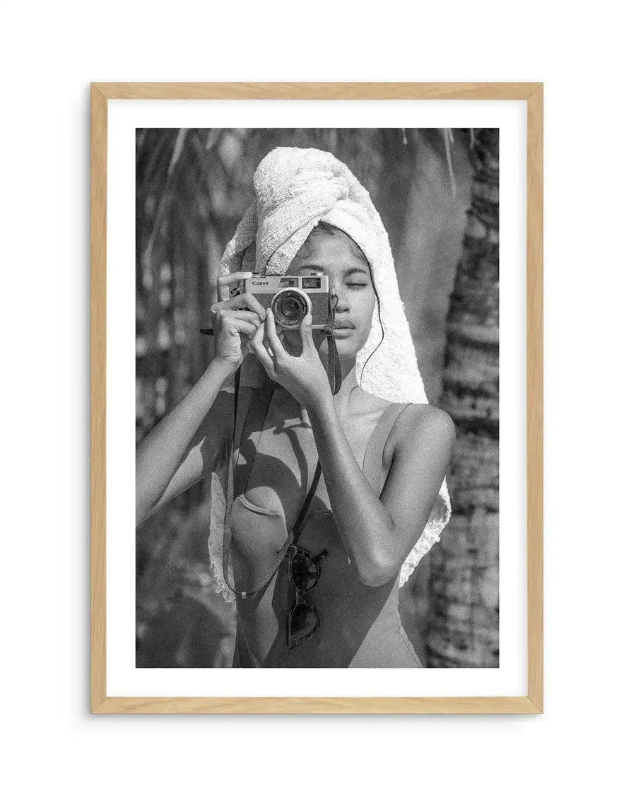 Framed black and white photograph of a young woman taking a picture with a vintage camera, displayed in a natural oak frame with white border. The woman, wearing a towel wrapped around her head and a swimsuit, holds a Canon camera up to her face, with her eyes closed, against a blurred background of palm trees.