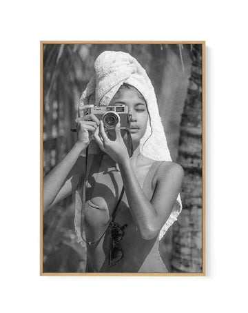 Framed black and white photograph of a young woman with a towel wrapped around her head, holding a vintage Canon camera up to her face as if taking a picture. She is wearing a swimsuit, and sunglasses hang from a strap around her neck. The background features blurred palm trees, suggesting a tropical outdoor setting, with a natural wood frame and white border.