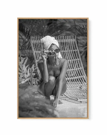 Framed black and white canvas print of a woman in a bikini and towel on her head, crouching on a beach with a camera. The woman is wearing sunglasses and holding a vintage camera up to her face, looking directly at the viewer. Behind her is a woven beach chair and palm trees. The print has a natural wood frame.