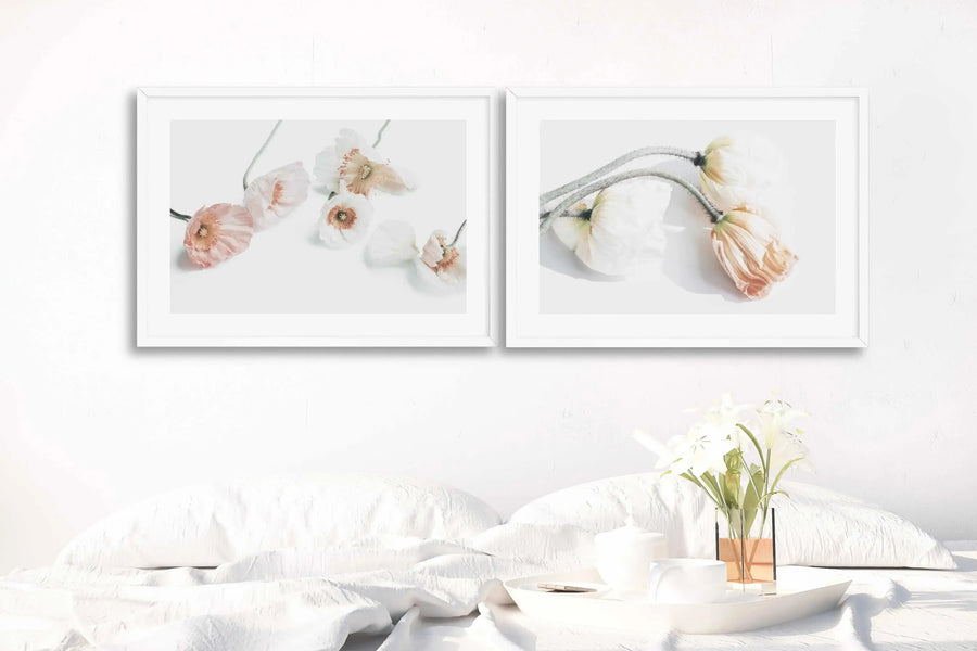 Lifestyle shot of two framed art prints displayed above a bed with white bedding and a white headboard. The left print features several light pink and white poppy flowers with delicate petals and visible stamens, arranged on a white background. The right print shows three poppy flowers, two white and one light orange, with their stems curving downwards, also on a white background. A white tray with a white cup, a lidded jar, and a vase with white flowers sits on the bed to the right.